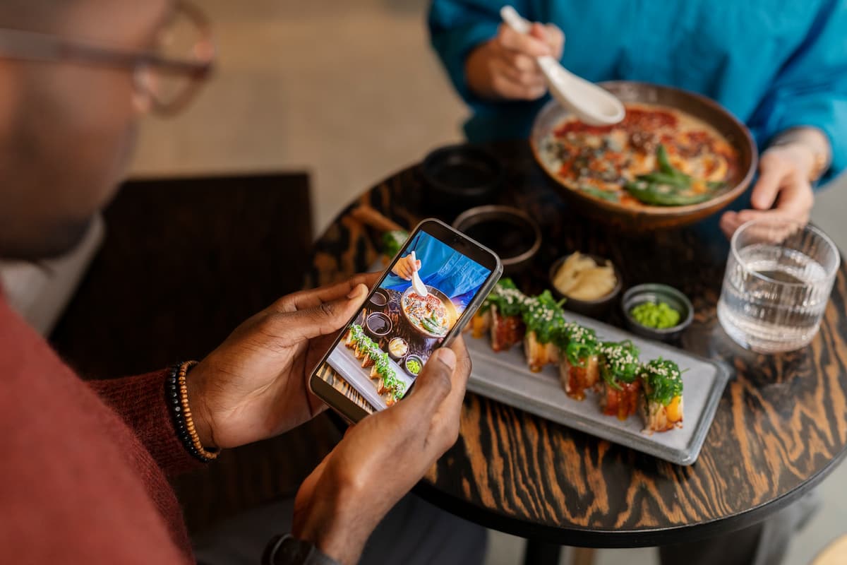 Hombre tomando una foto con su teléfono de un plato de comida rápida gourmet en un restaurante, junto a una bandeja de sushi.