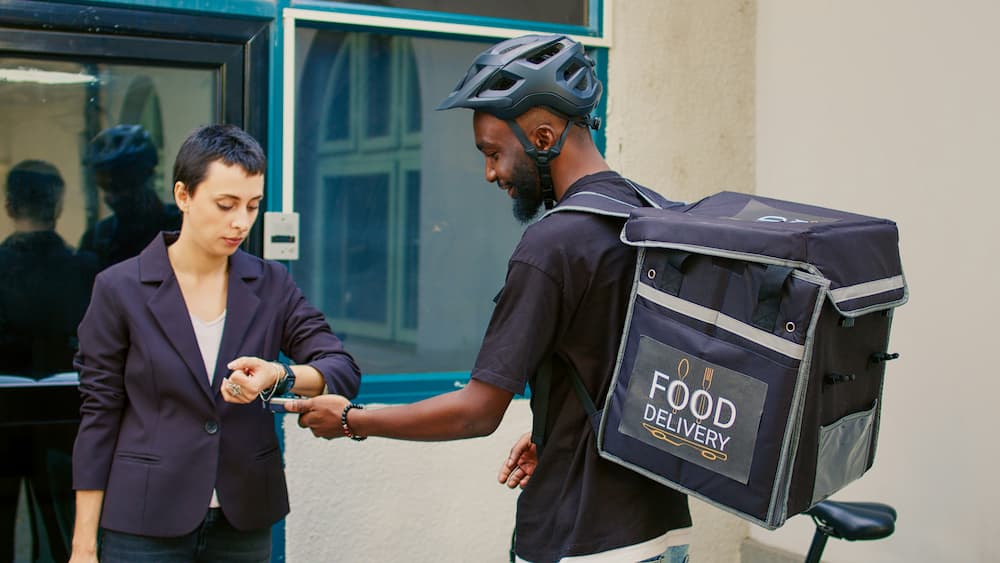 Entrega de comida con repartidor usando mochila de delivery. Una mujer espera el pedido.