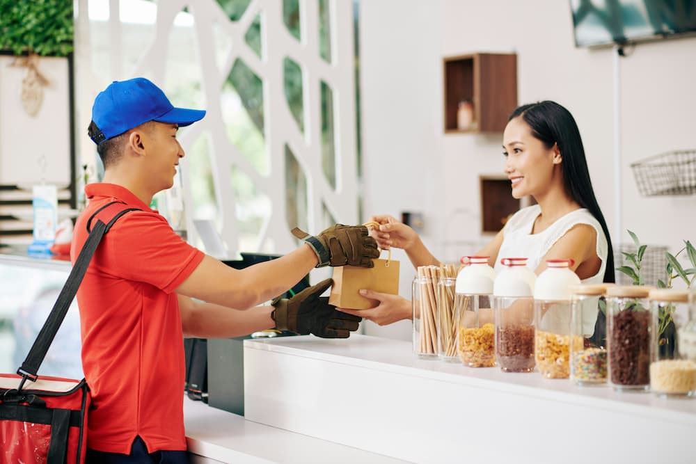 Repartidor recibiendo un pedido en una cafetería moderna, interactuando con una empleada que sonríe mientras entrega los productos