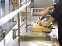 La imagen muestra una cocina profesional con encimeras de acero inoxidable. Un chef, vestido con un uniforme negro, está cortando una verdura verde (parece un puerro) sobre una tabla de madera. En primer plano se ve un fregadero metálico, y al fondo hay cuencos con verduras y utensilios de cocina.