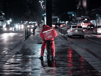 La imagen muestra a un repartidor nocturno de la plataforma Rappi, vestido con uniforme rojo y utilizando una bicicleta como medio de transporte. Lleva una mochila térmica grande para entregas, claramente visible con el logotipo de la empresa.