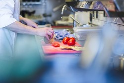 Una persona trabajando en una dark kitchen profesional, cortando un vegetal en una tabla de cortar roja. En la tabla también hay tomates y lo que parece ser pan. La cocina está equipada con utensilios, una estufa y recipientes de almacenamiento, representando el ambiente dinámico y práctico de este tipo de cocinas destinadas a la entrega de comida a domicilio.