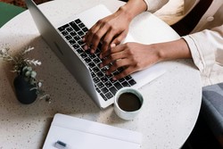 Una persona escribe en una computadora portátil que está sobre una mesa redonda de madera clara. En la mesa, también hay una taza de café, una libreta blanca con una pluma y un pequeño florero negro con flores, creando una atmósfera de trabajo relajada y bien organizada