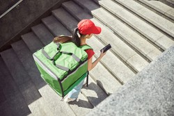 Una persona con gorra roja y camiseta roja sube unas escaleras llevando una mochila verde grande en la espalda, típica de repartidores. En su mano derecha sostiene un teléfono móvil, mientras parece concentrada en su labor diaria.