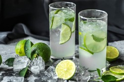 Dos vasos altos llenos de una bebida refrescante, adornada con rodajas de lima y hojas frescas de menta. Están rodeados de cubos de hielo y limas frescas, descansando sobre una superficie de mármol gris.