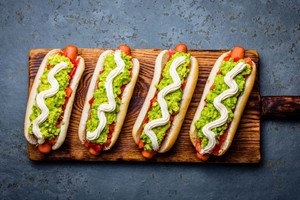 Cuatro completos con palta, tomate y mayonesa sobre tabla de madera, representando tipos de completos chilenos tradicionales.