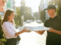La imagen muestra a una mujer recibiendo cajas de pizza de un repartidor en una calle soleada. Ambos están sonriendo de manera amistosa.