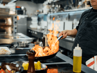 Se ve a un chef o cocinero preparando un plato. La acción principal capturada es el flameado (o flambé), donde se añade un licor caliente a la sartén.