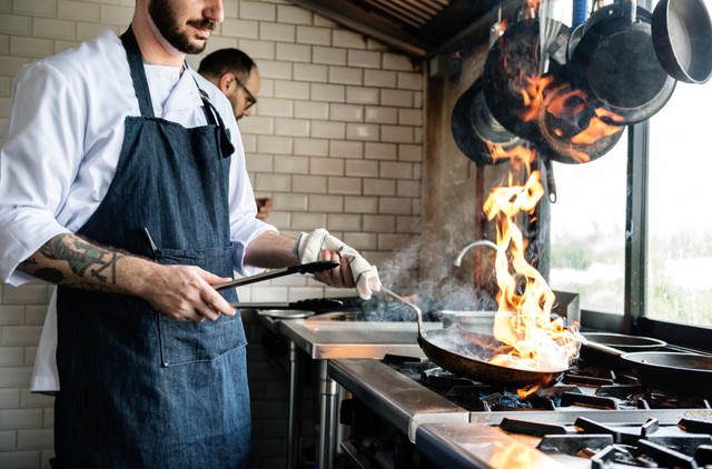 dark kitchen: Hombre flanqueando la cazuela lleva un delantal azul en una cocina industrial, hay llamas saliendo de la estufa