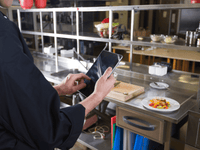 La imagen es un plano medio que muestra a un chef o cocinero, vestido con una chaqueta de chef (filipina) negra, interactuando con una tableta digital en una cocina de restaurante profesional.