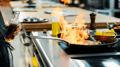 La imagen muestra a un cocinero preparando un plato en una cocina profesional, con una sartén sobre el fuego produciendo una gran llamarada (flambear).