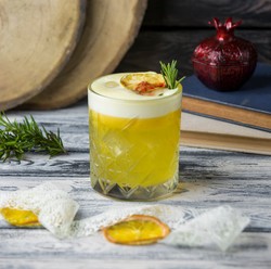 Un vaso de cristal tallado con una bebida espumosa de color amarillo, decorado con una rodaja de limón seco y una ramita de romero. La bebida descansa sobre una mesa de madera clara junto a ramitas de romero y rodajas de limón seco.