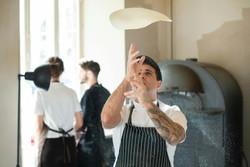 Un chef tatuado en el brazo lanza una masa de pizza al aire con precisión y destreza en una cocina profesional. Al fondo, se ven dos personas más trabajando, una de ellas de espaldas, añadiendo movimiento y dinamismo a la escena