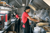 Restaurantes en casa: Un hombre y una mujer trabajando uno al lado del otro, ambos con delantales rojos y negros y una gorra, preparando comida en la plancha, en una cocina pequeña pero bien equipada.