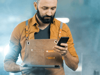 La imagen muestra a un hombre con barba y cabello oscuros, vestido con una camisa de manga larga de color mostaza o marrón claro y un delantal de trabajo de cuero o lona color caqui con detalles de cuero. Está mirando la pantalla de un smartphone que sostiene en su mano derecha, mientras que en la mano izquierda sujeta una pequeña carpeta o tableta delgada.
