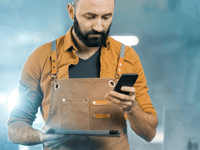 La imagen muestra a un hombre con barba y cabello oscuros, vestido con una camisa de manga larga de color mostaza o marrón claro y un delantal de trabajo de cuero o lona color caqui con detalles de cuero. Está mirando la pantalla de un smartphone que sostiene en su mano derecha, mientras que en la mano izquierda sujeta una pequeña carpeta o tableta delgada.