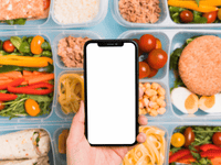 La imagen muestra una vista aérea (plano cenital) de una mano sosteniendo un smartphone con la pantalla en blanco, enmarcado por una variedad de recipientes de comida preparada, comúnmente conocidos como meal prep o tuppers.