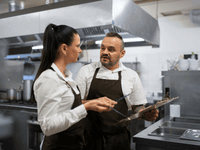 En la imagen se observa a dos trabajadores de una cocina industrial conversando mientras revisan una lista de control.